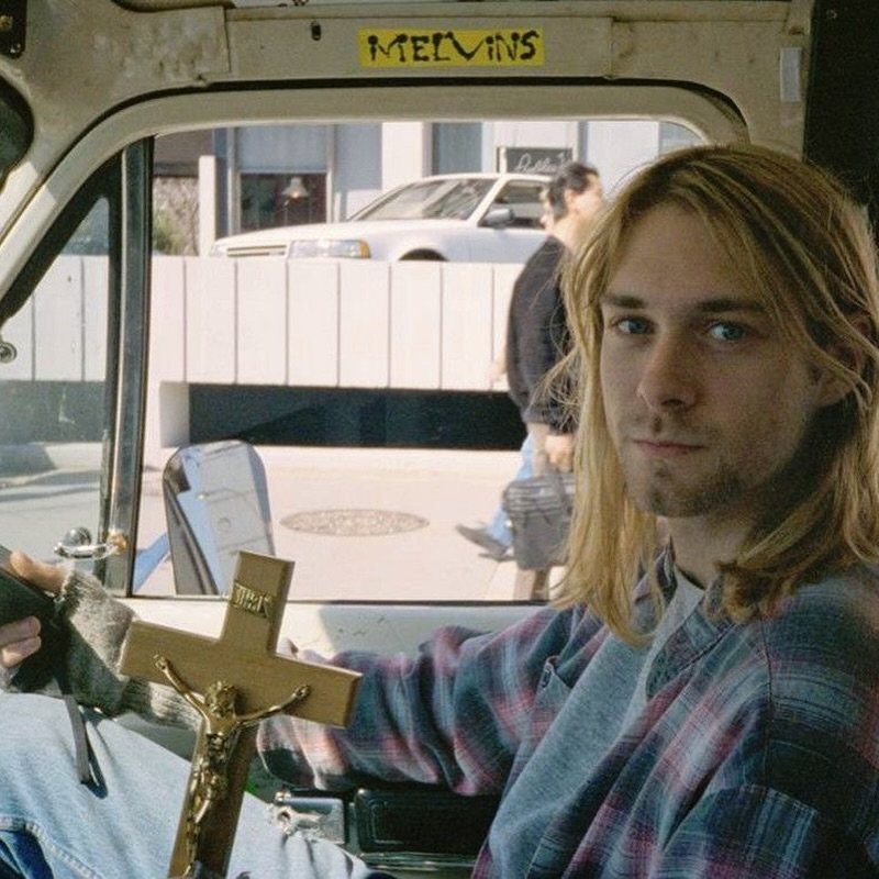 A young man with long blonde grunge waves sitting in a vehicle holding a crucifix.
