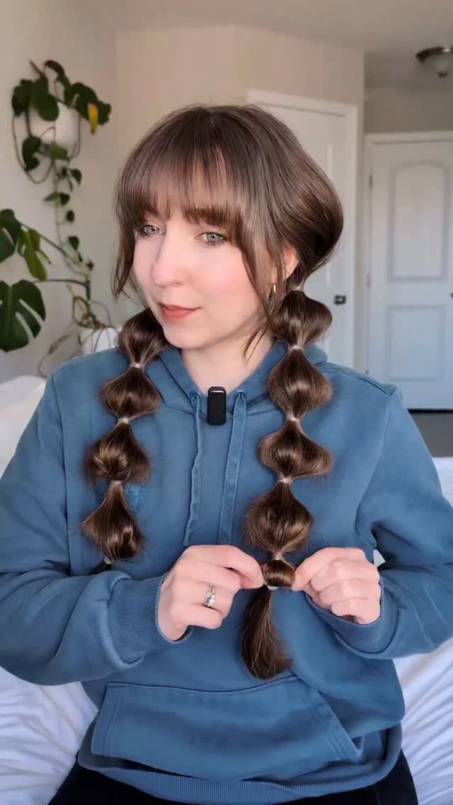 A woman styling her brown hair into two textured bubble braids with elastic bands.