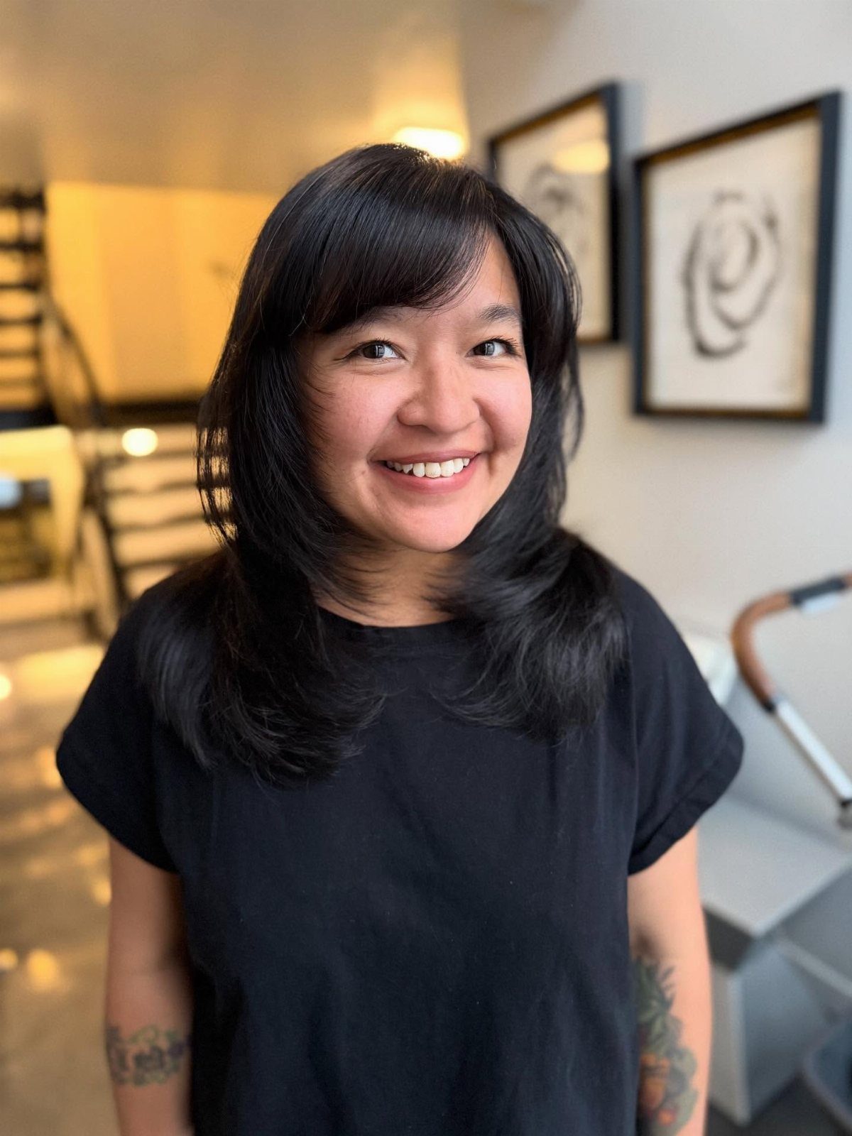 A smiling woman with layered black hair and side-swept bangs wearing a black t-shirt.