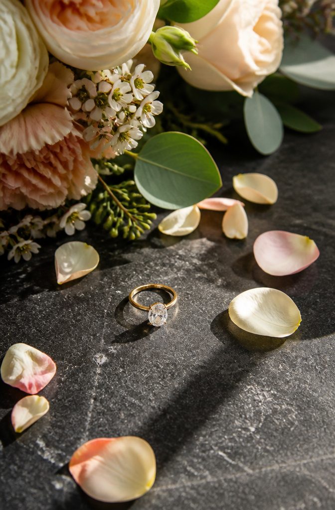 An engagement ring on a dark surface with flower petals around it.