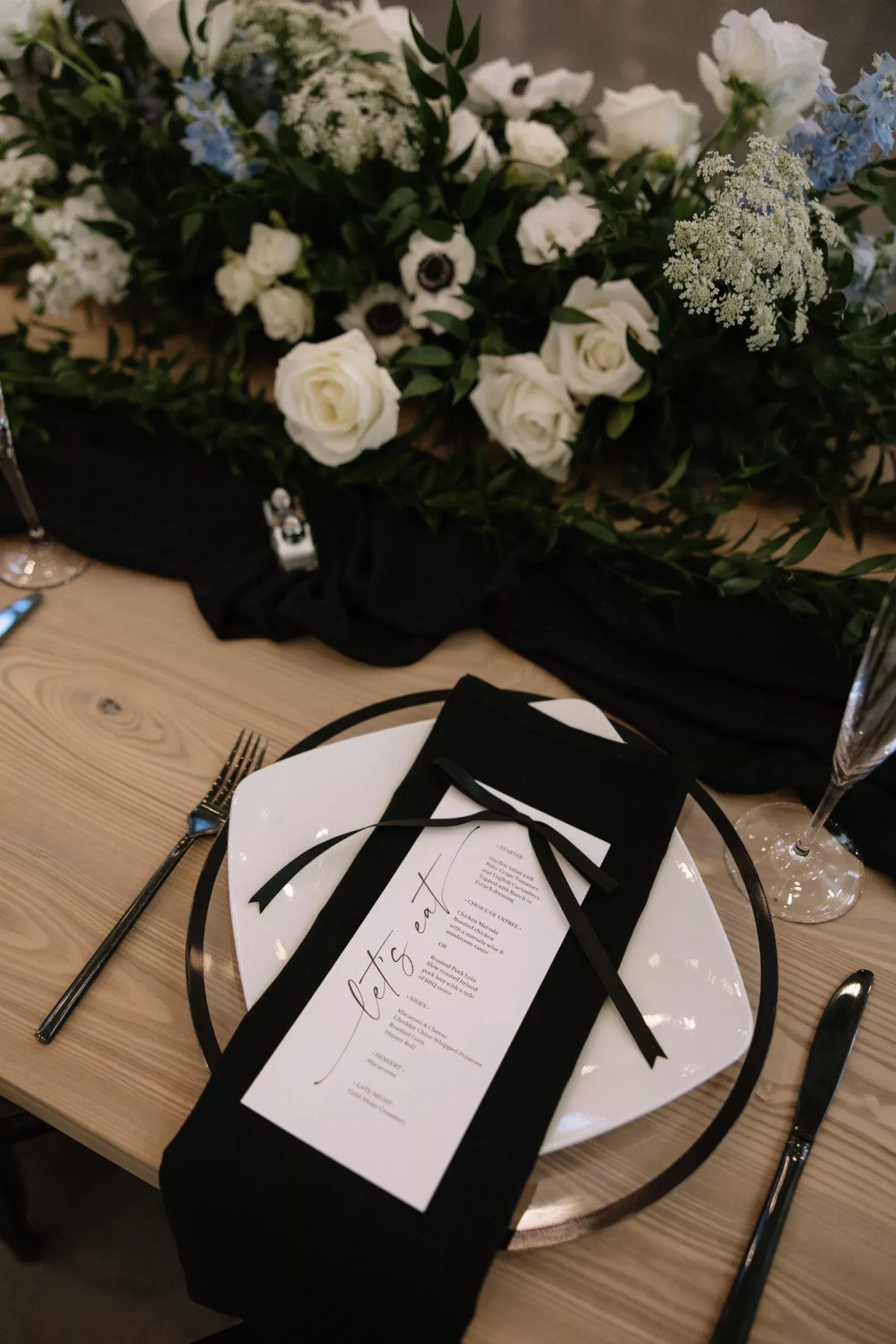 White floral arrangement with greenery and roses on a black table runner with place settings.