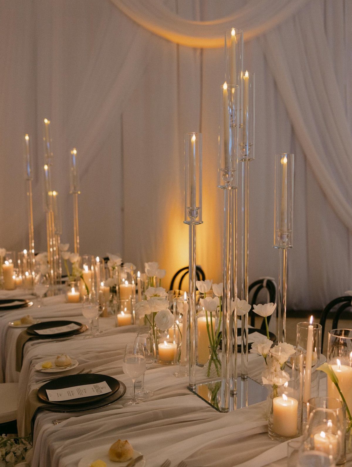 Tall glass candle holders with lit white pillar candles on a white wedding table.