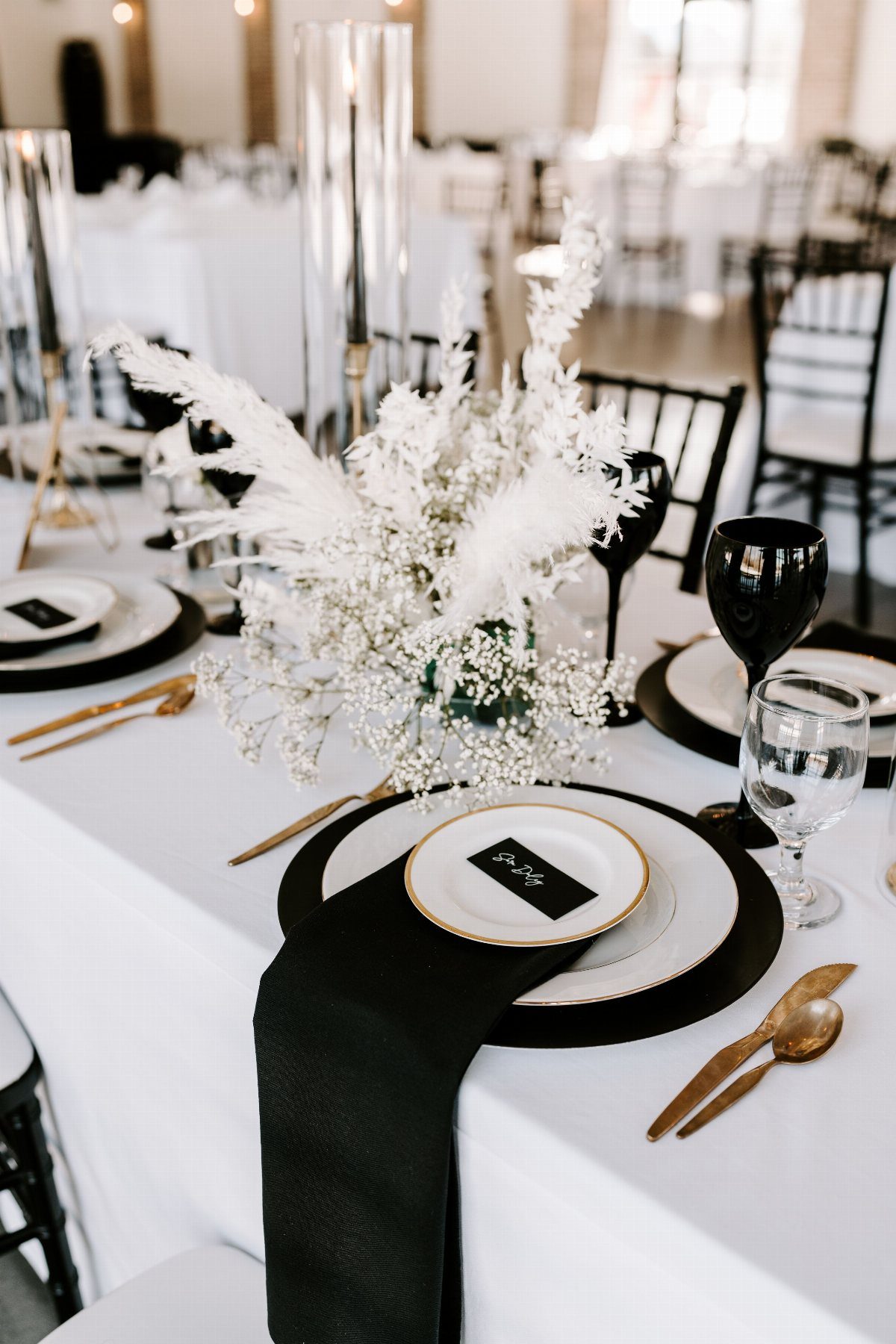 Wedding table setting featuring white plates, black chargers, black napkins, and gold cutlery with white flowers.