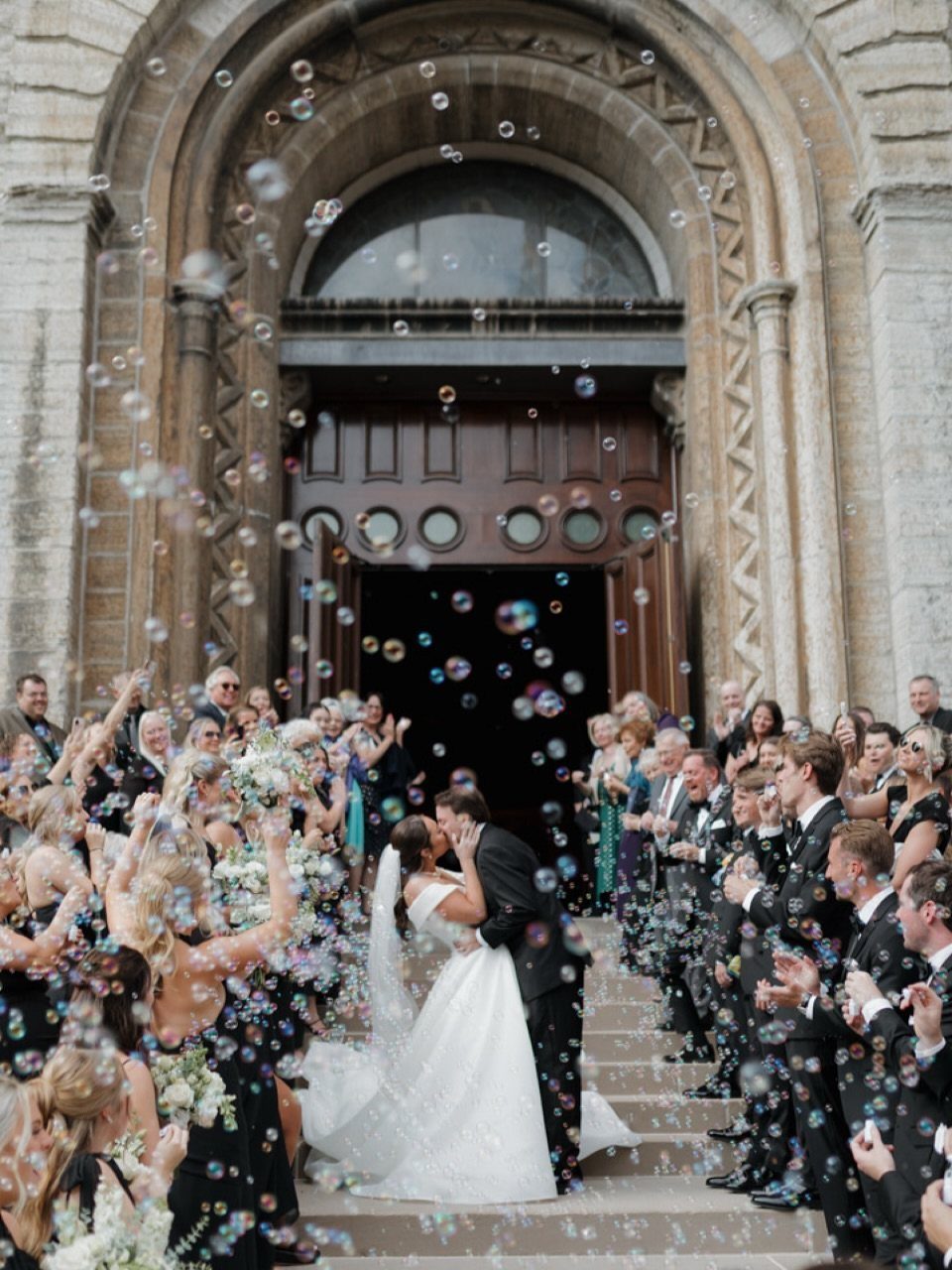 Bride and groom kissing on church steps surrounded by guests blowing bubbles.
