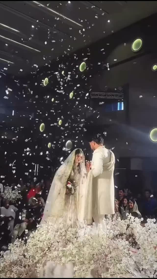 A bride and groom stand together under falling white confetti surrounded by white flowers.