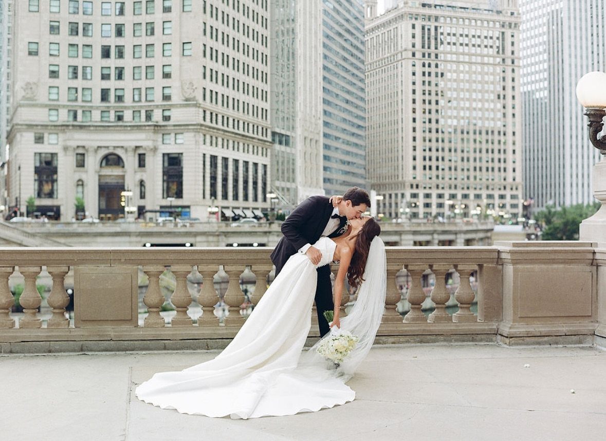 A bride and groom kissing while the groom performs a romantic dip on a city bridge.