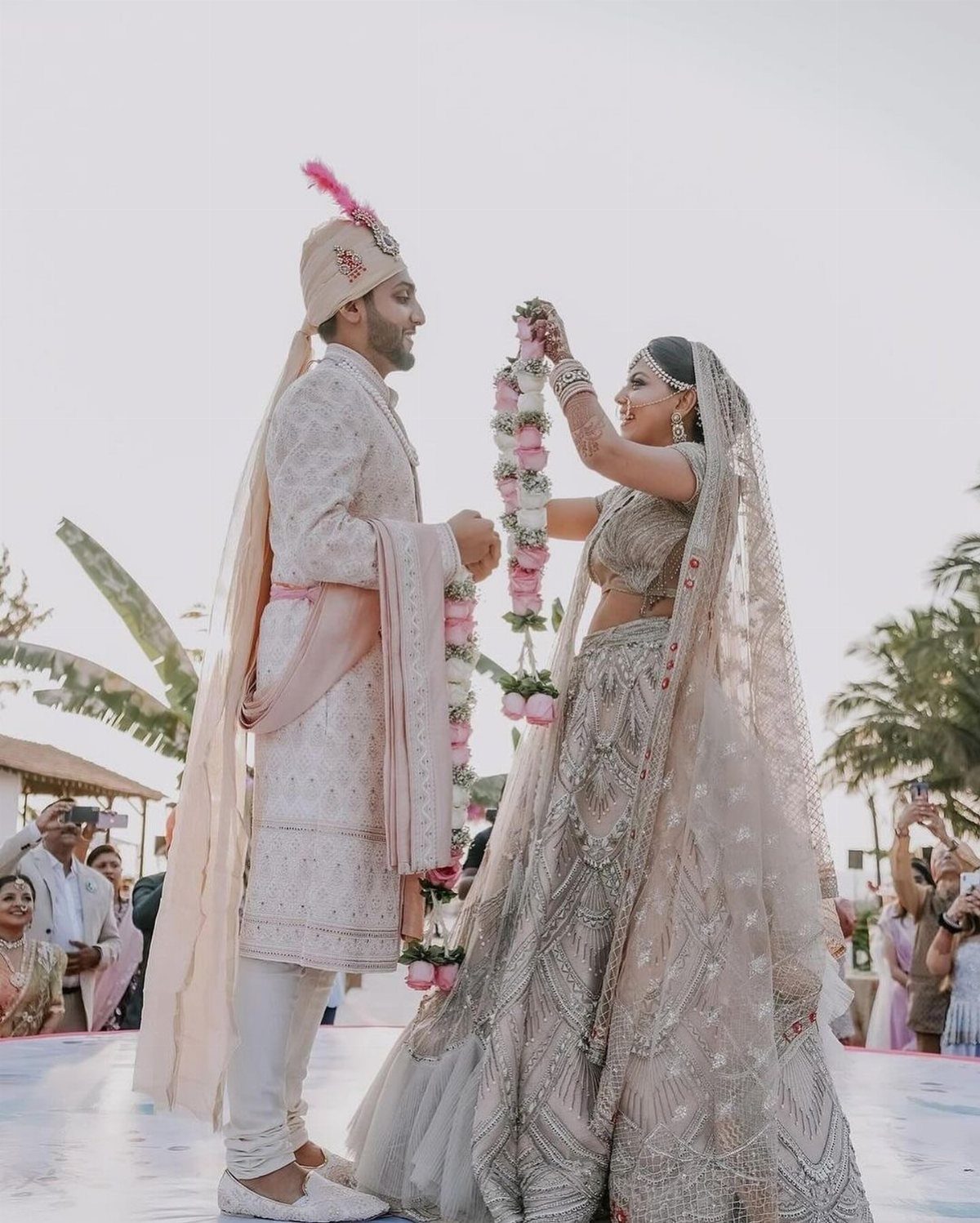 A bride and groom in traditional Indian wedding attire facing each other outdoors.