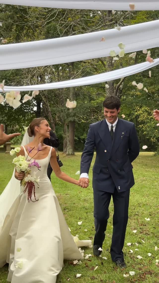 A bride and groom walk hand in hand through falling flower petals outdoors.