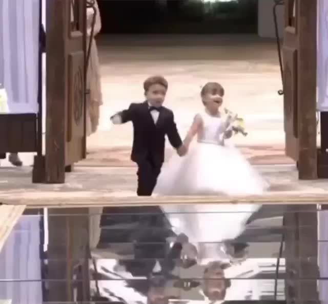 Two children in formal wedding attire running joyfully through an open doorway while holding hands.