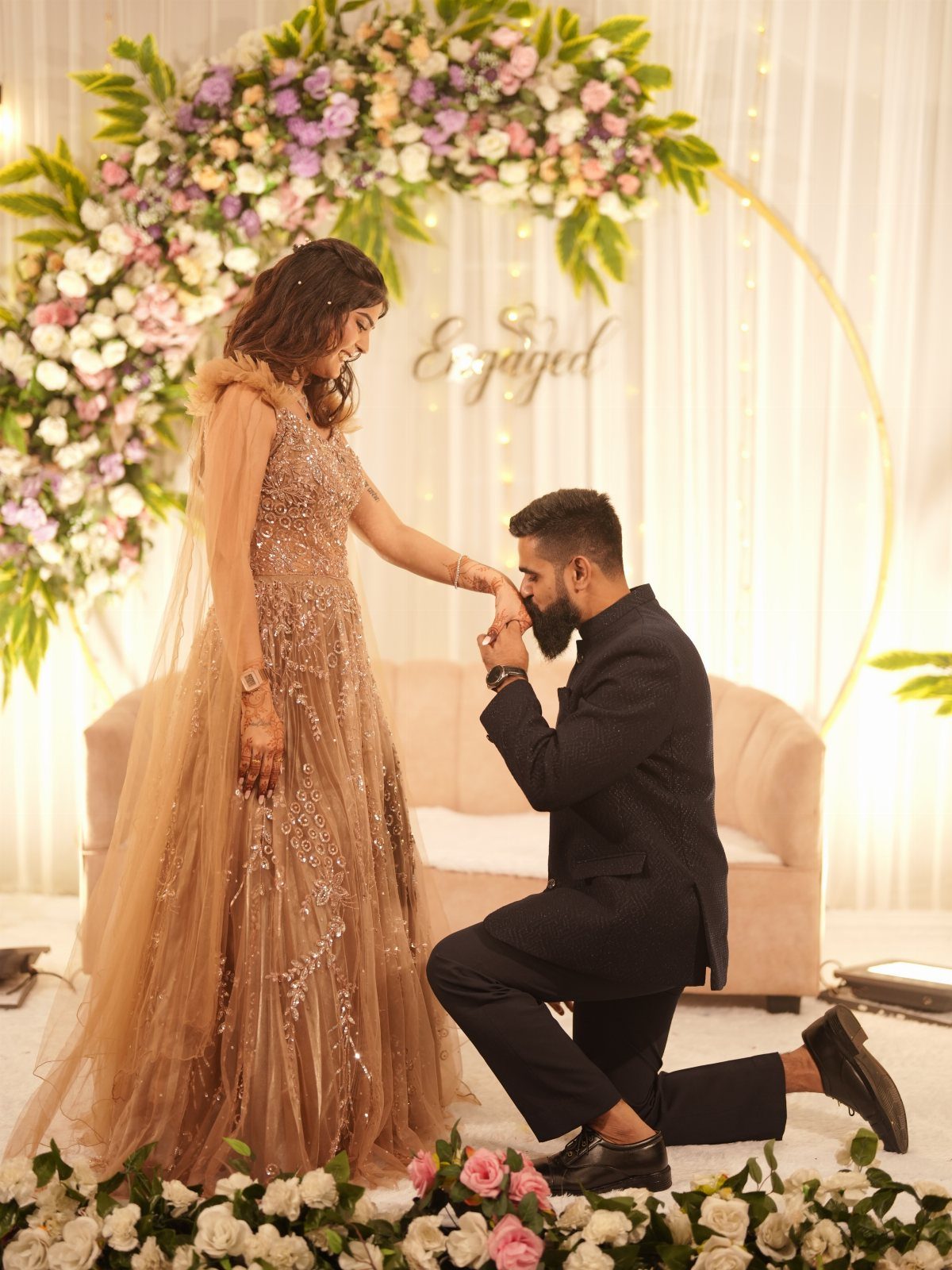 A man in a suit kneels to kiss the hand of a woman in a sparkling gown.