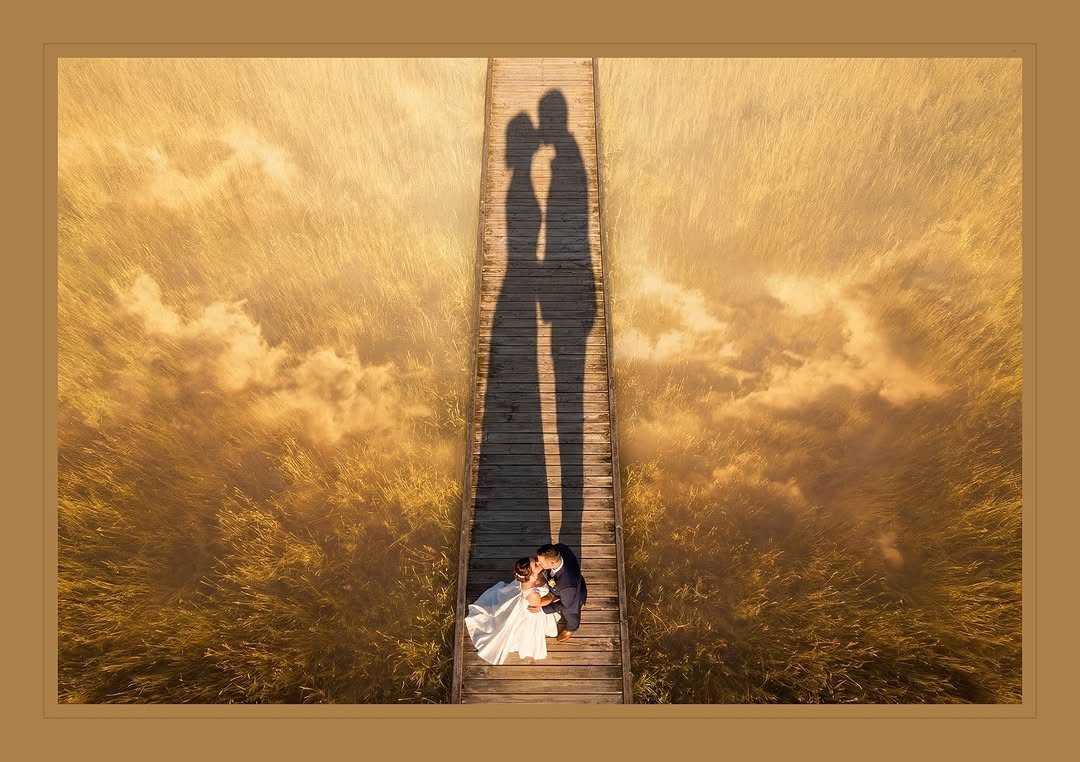 A bride and groom stand on a wooden path surrounded by golden grass with long shadows.