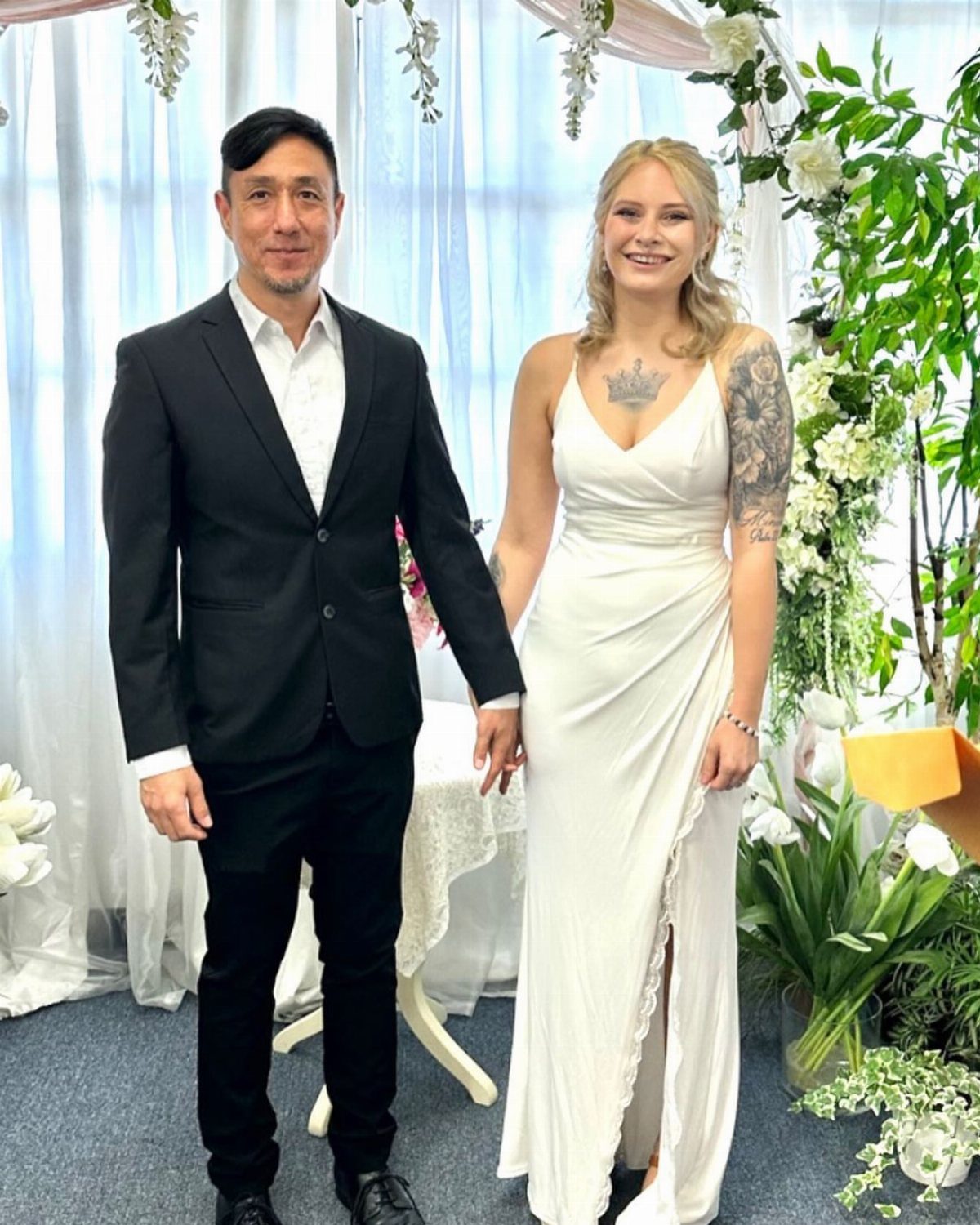 A bride and groom in wedding attire smiling and holding hands indoors.