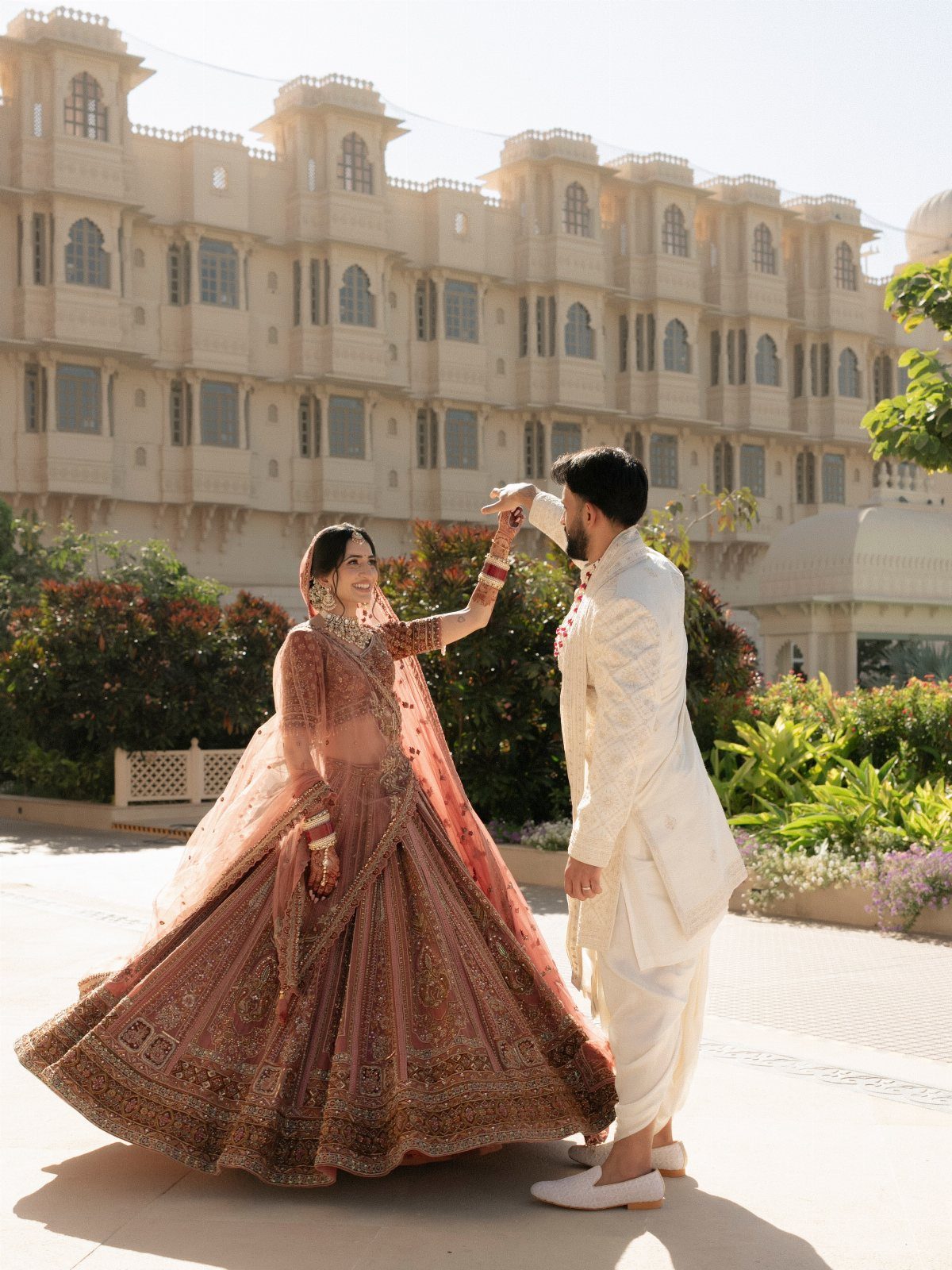 Bride in a pink lehenga twirls while holding hands with her groom outdoors.