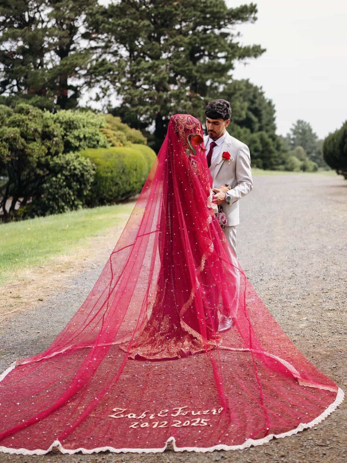 A bride wearing a long, flowing red veil that spreads across a gravel path next to her groom.