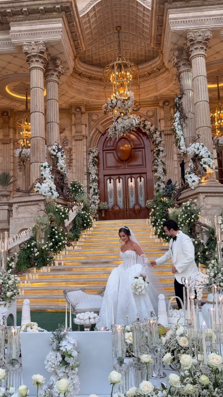 Bride and groom standing on a grand, flower-decorated staircase in an ornate hall.