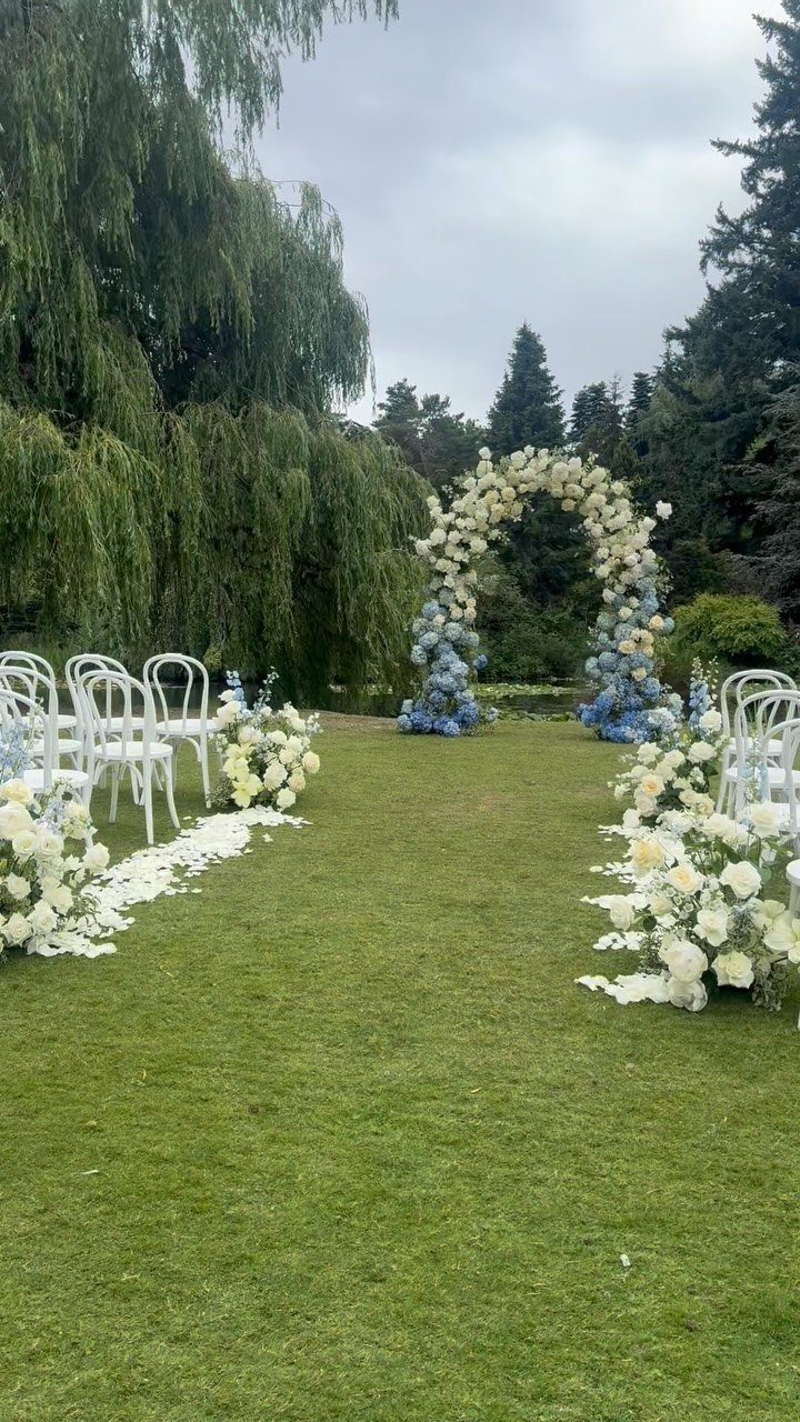 A wedding ceremony aisle with white chairs, flower arrangements, and a floral arch on a green lawn.