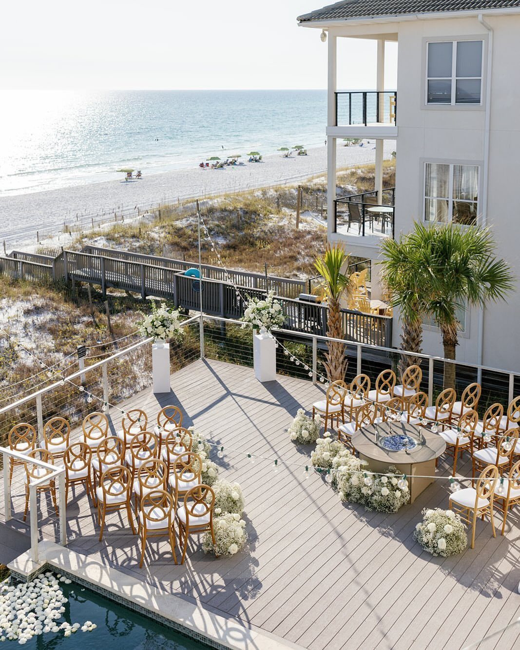 Wedding ceremony setup with gold chairs on a wooden deck overlooking a beach and ocean.