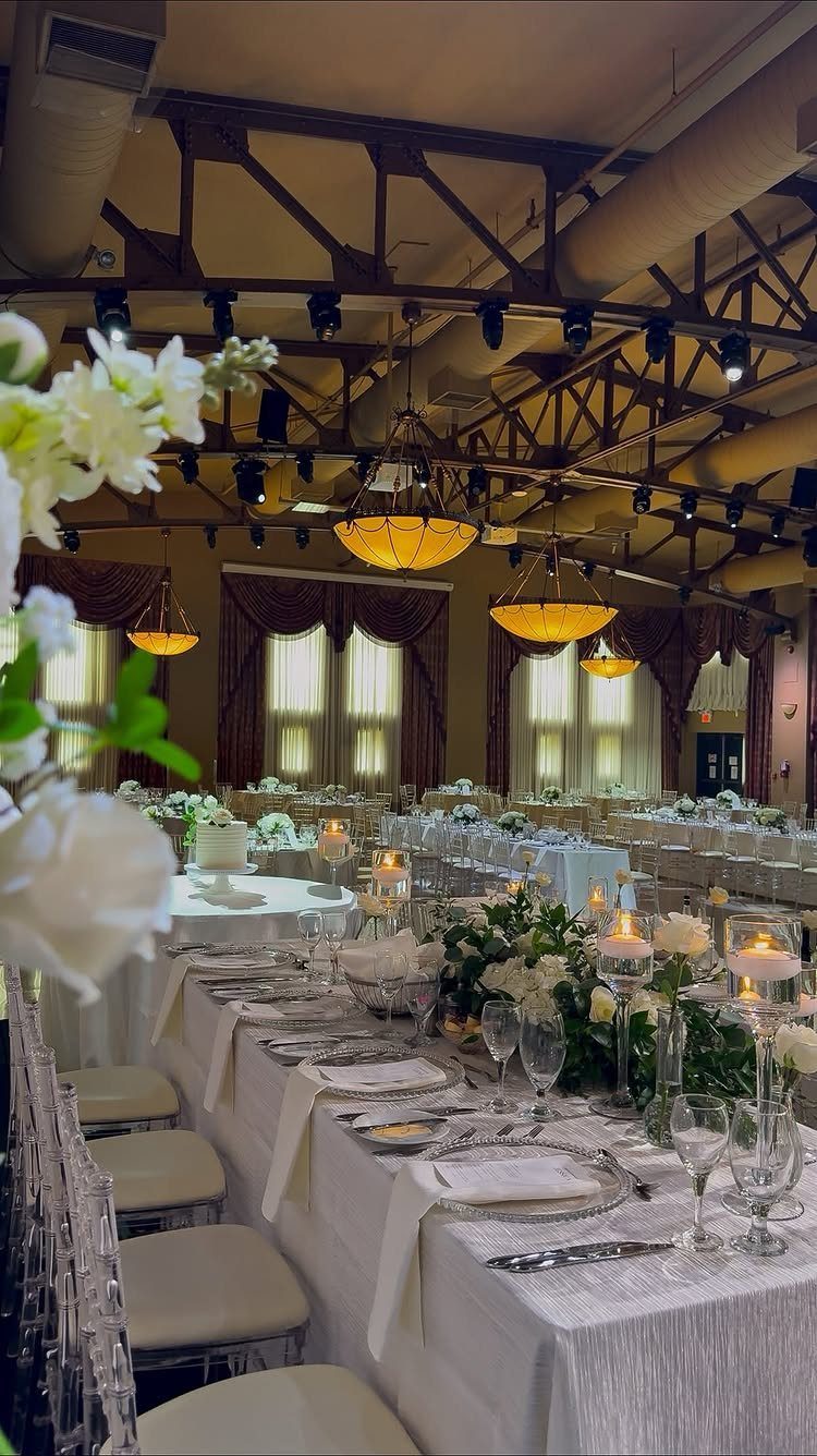 A formal wedding reception hall with white tablecloths, floral centerpieces, and large amber chandeliers.