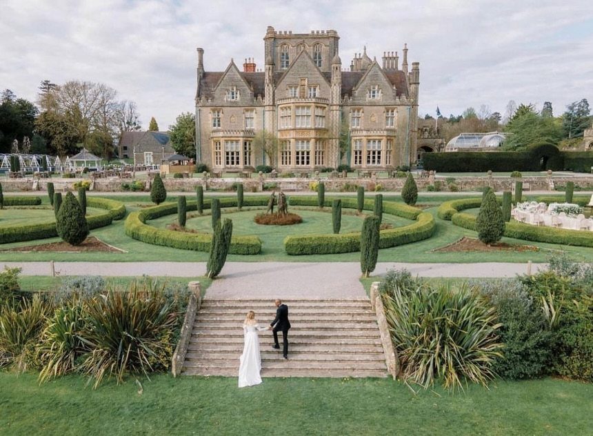 A bride and groom walk down stone stairs toward a grand Victorian mansion and formal gardens.