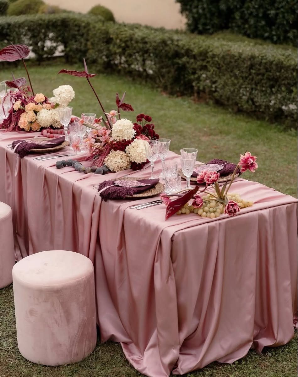 A long table set with blush pink satin linens and burgundy floral arrangements outdoors.