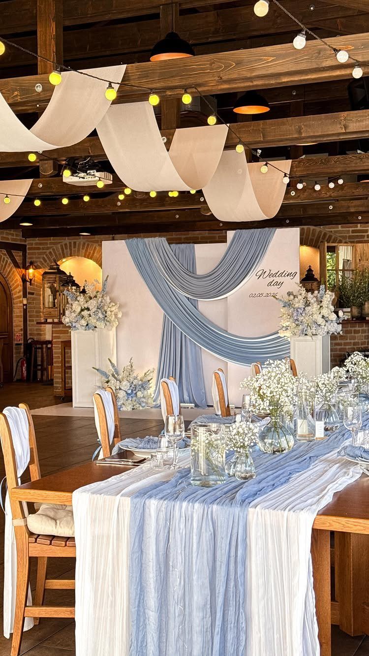 A wedding table decorated with light blue linens, white flowers, and glass vases under string lights.