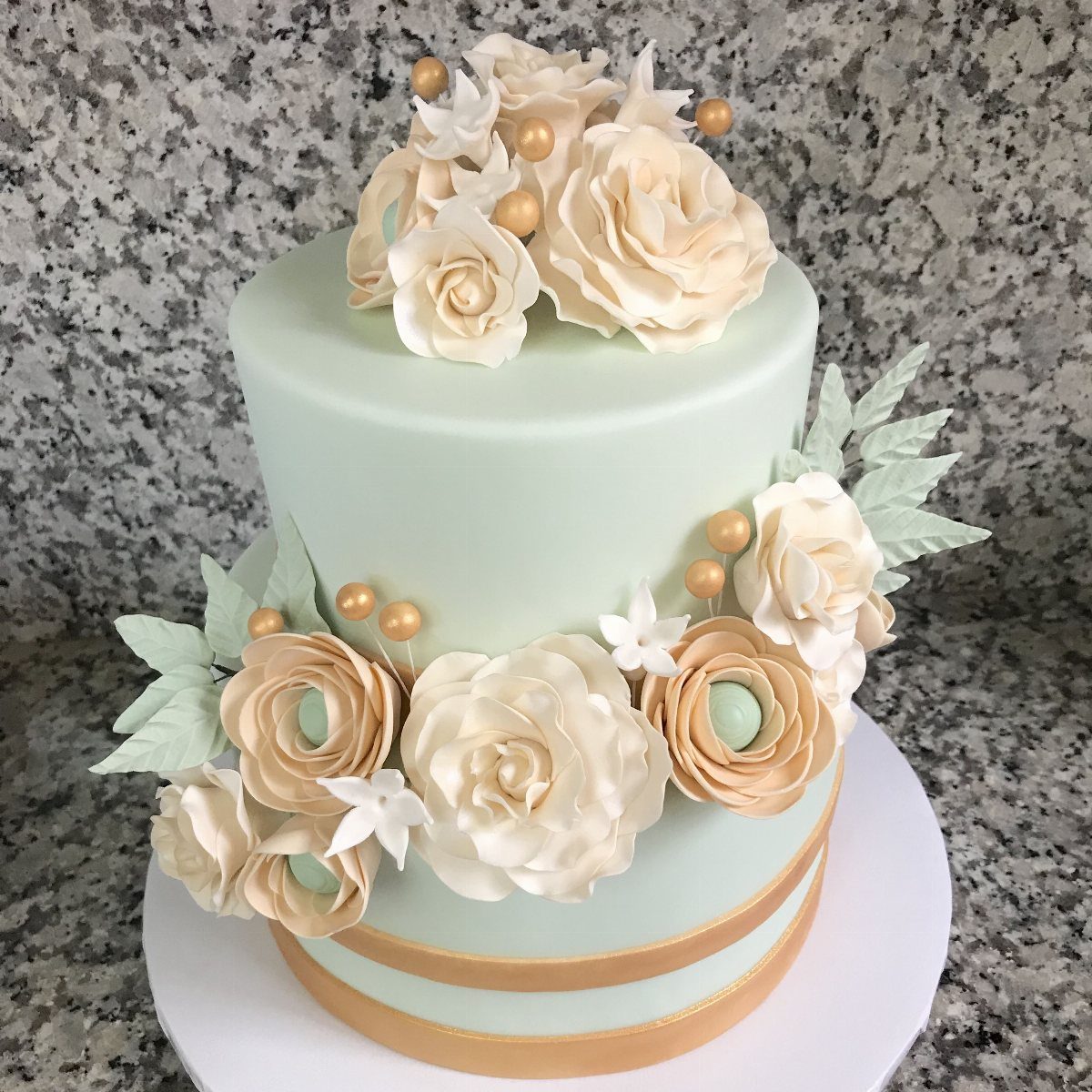 A two-tiered mint green cake adorned with cream sugar flowers and gold accents on granite.