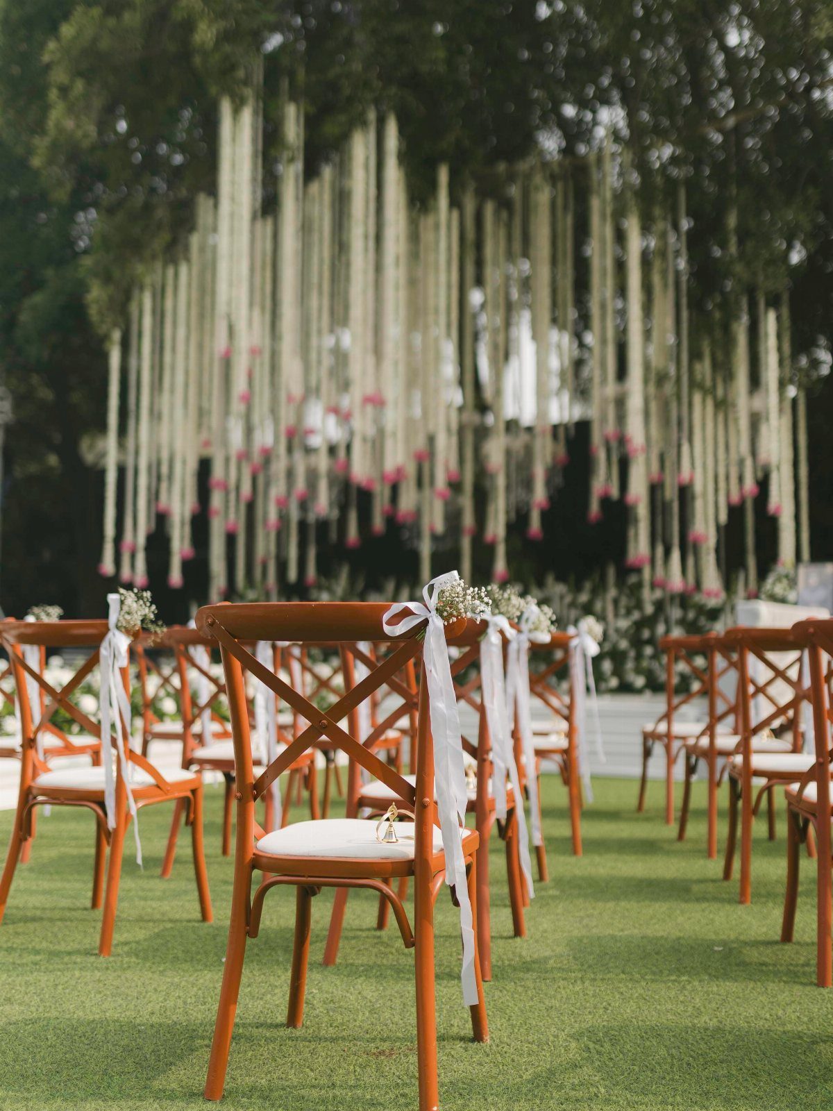 Wooden cross-back chairs decorated with white ribbons and flowers arranged on green grass.