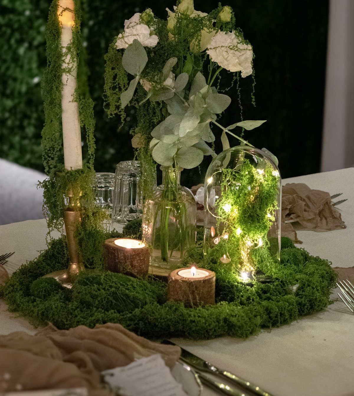 A moss-covered centerpiece with a fairy light terrarium, white flowers, and a tall candle.