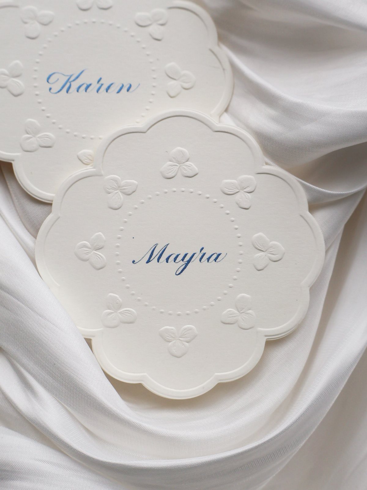 Two white scalloped place cards with blue calligraphy names resting on white fabric.