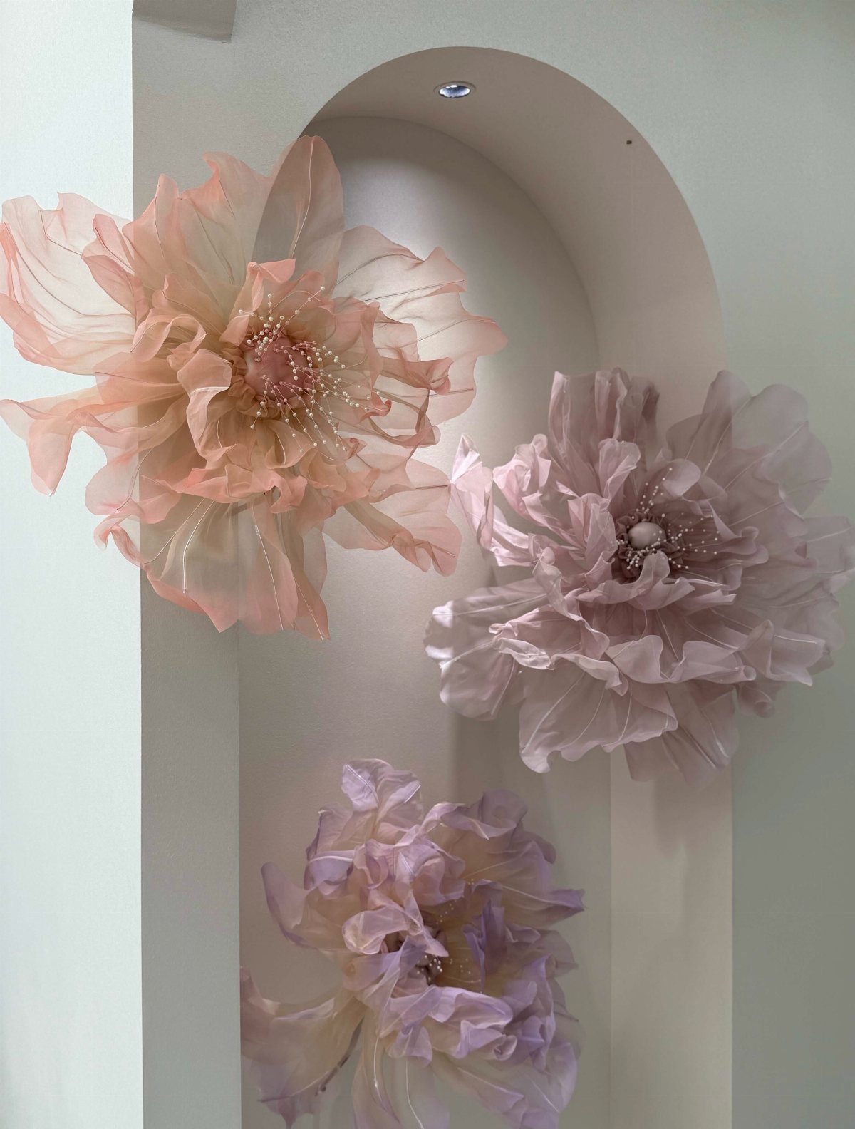 Three large, ruffled fabric flowers in peach and lavender tones inside a white arched niche.