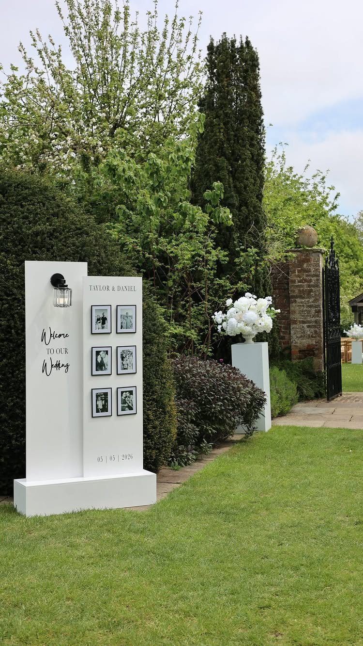 A large white wedding welcome sign with photographs and calligraphy on a green lawn.