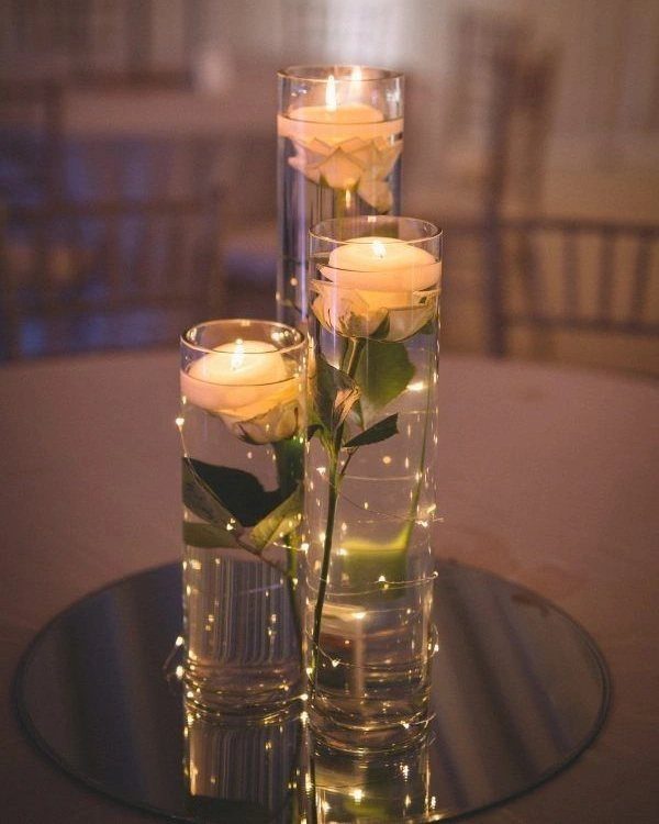 Three tall glass vases with floating candles and white roses on a mirrored table.