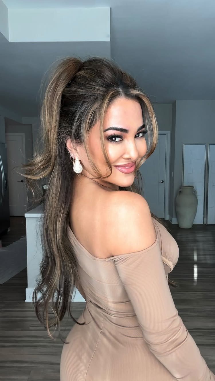 A woman with long wavy hair in a high ponytail posing in a beige dress.
