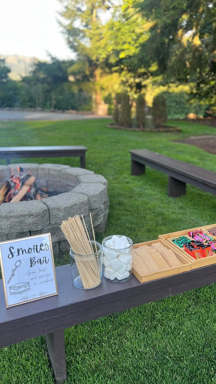 A s'mores bar setup with skewers, marshmallows, and candy on a wooden table near a fire pit.