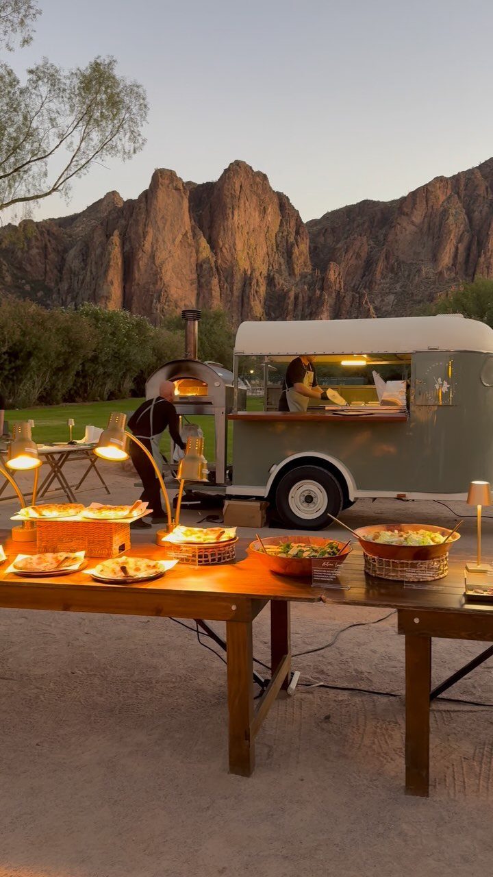 A catering trailer serving food on a wooden table outdoors against a mountain backdrop.