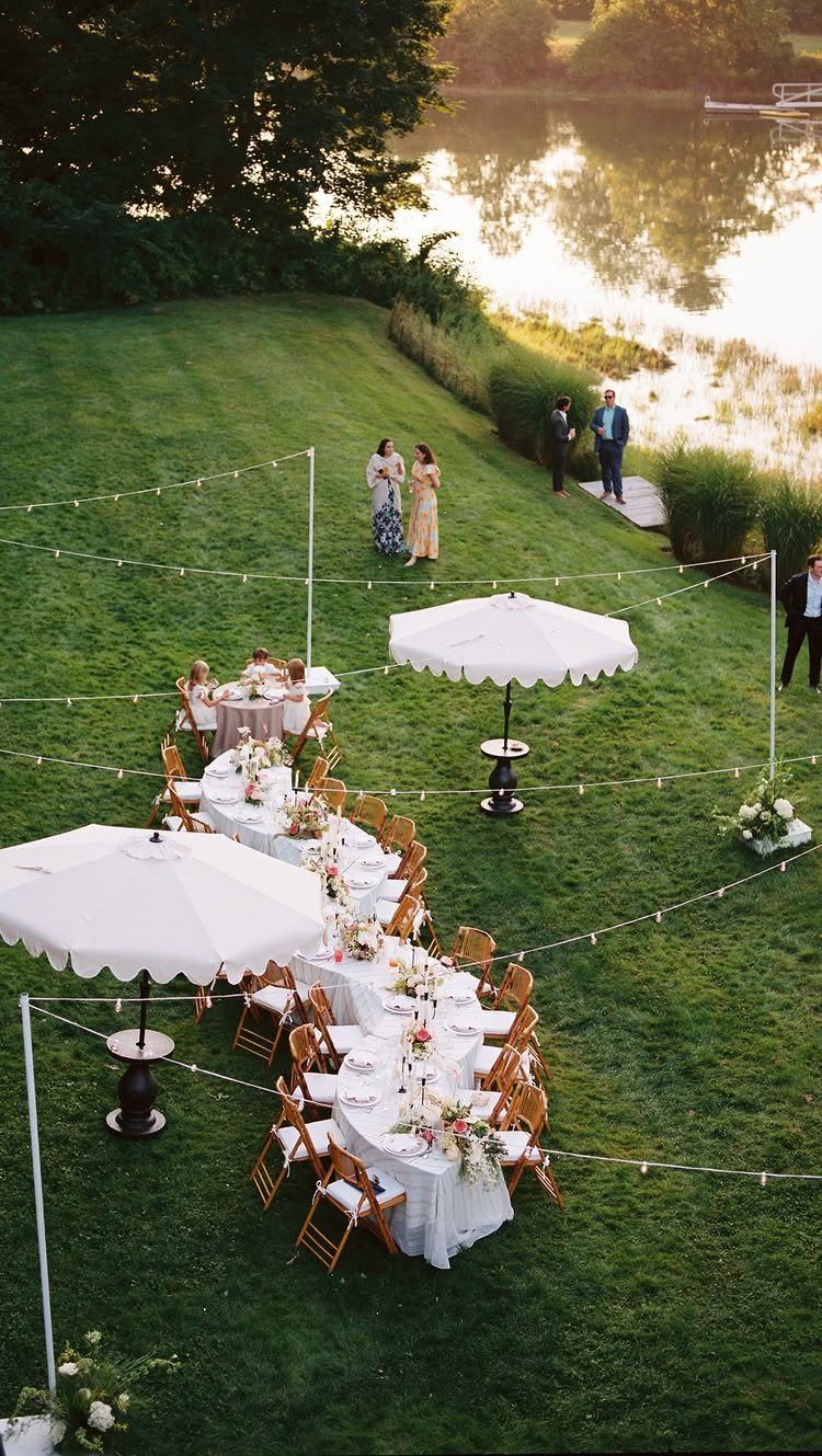 Long dining tables with white linens and floral arrangements set on a grassy lawn by a lake.