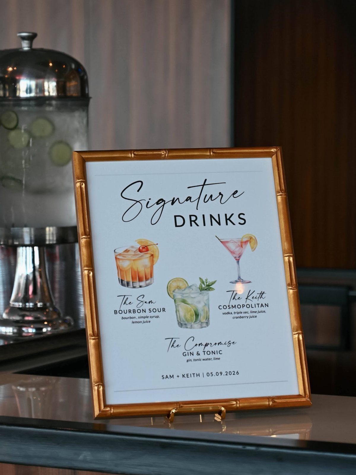 A gold framed sign displaying watercolor illustrations of signature wedding cocktails on a bar counter.