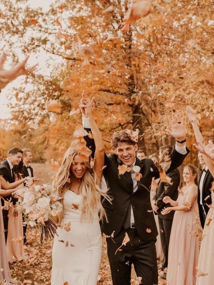 A happy bride and groom celebrating outdoors under falling autumn leaves surrounded by wedding guests.