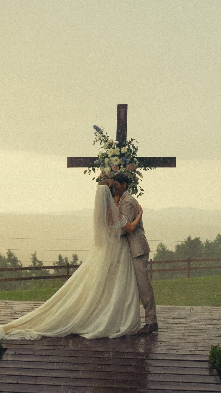 A bride and groom kissing behind a large, flower-decorated wooden cross in the rain.