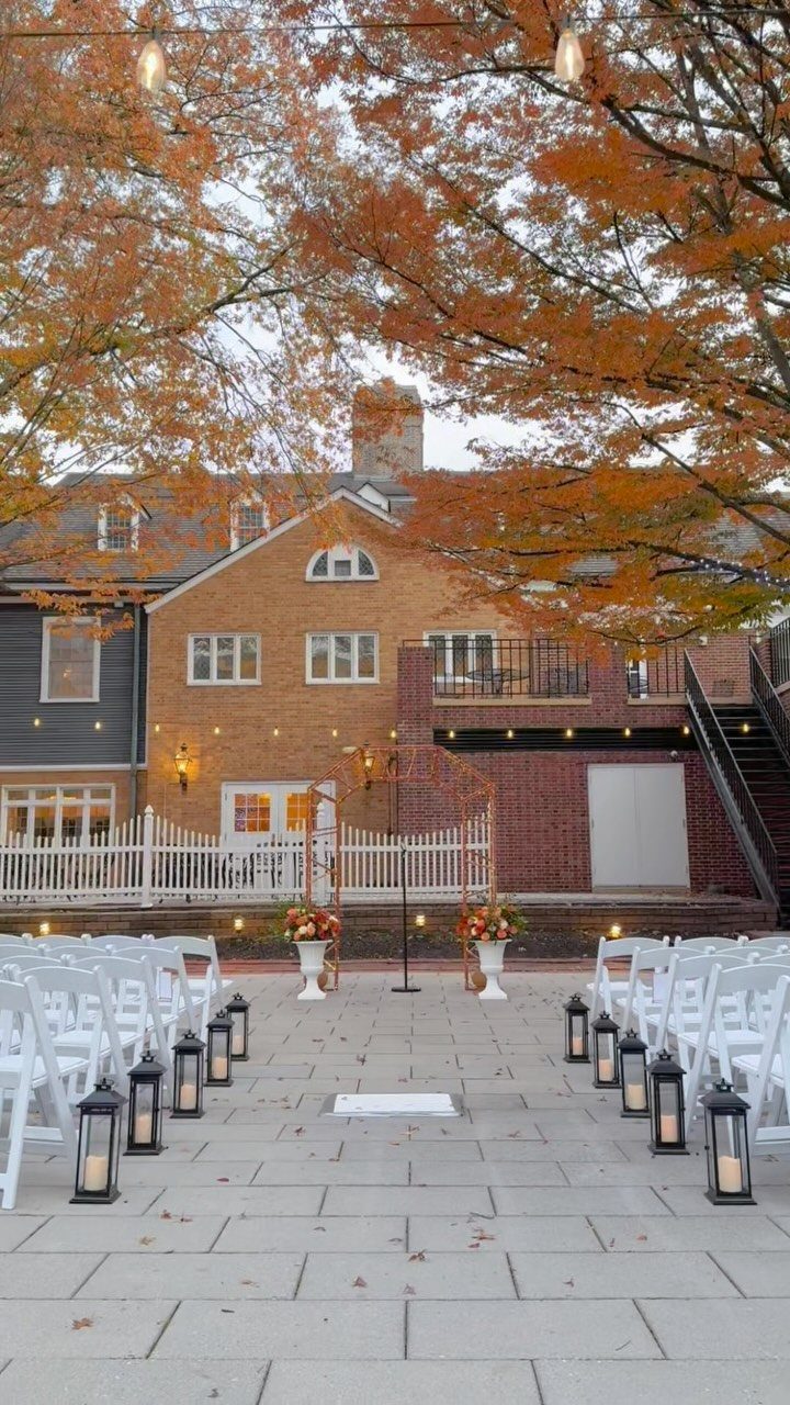 White wedding chairs arranged on a stone patio under orange autumn trees.