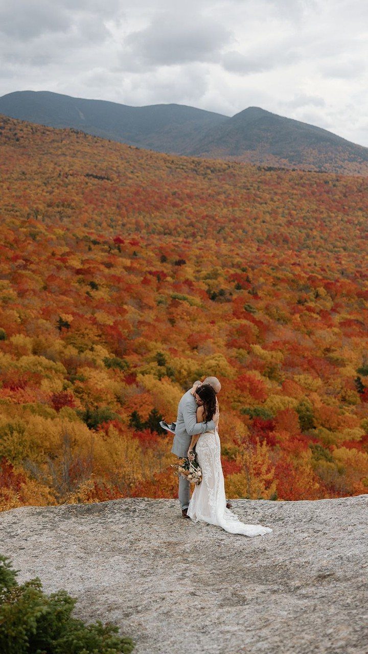 A bride and groom embrace on a rocky ledge overlooking a colorful autumn forest and mountains.