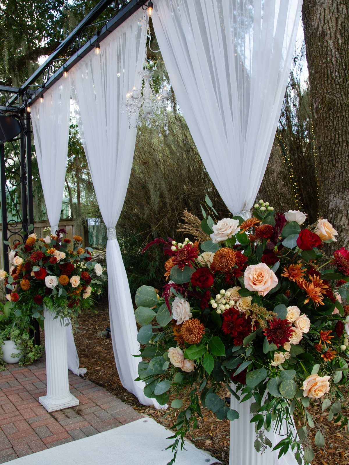 White draped fabric and large autumn-colored floral arrangements on pedestals at an outdoor wedding altar.