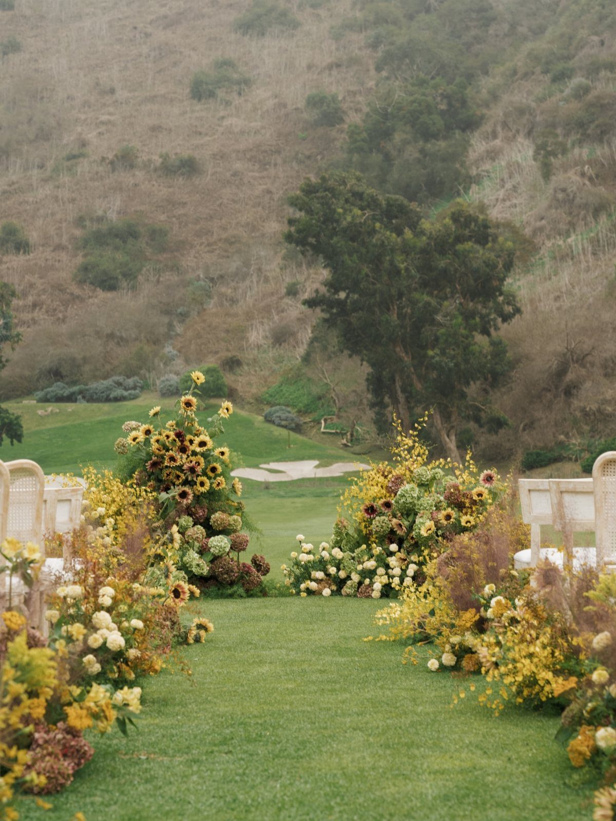 An outdoor wedding aisle decorated with sunflowers and autumnal floral arrangements on a green lawn.