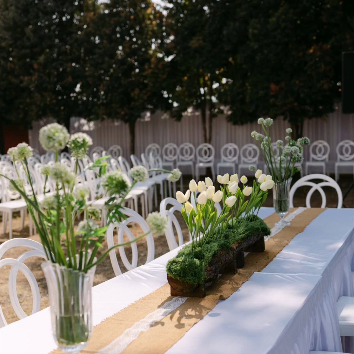 A long wedding table featuring a burlap runner, white tablecloth, and mossy tulip centerpiece.