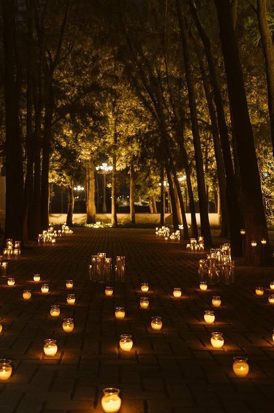 Small glowing glass candle lanterns scattered across a dark brick path between tall trees at night.