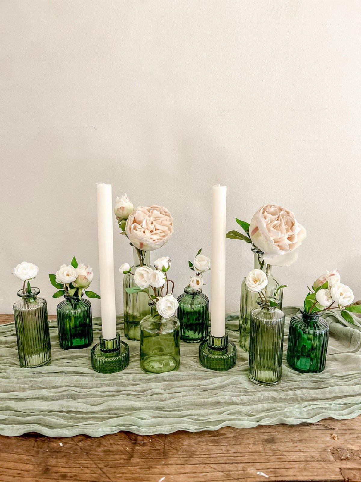 Green and clear glass vases with flowers and candles on a textured fabric runner.