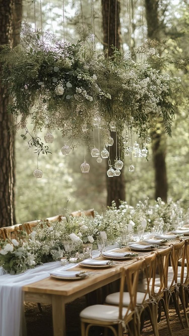 A long wooden wedding table decorated with white flowers, greenery, and a white fabric runner in a forest.