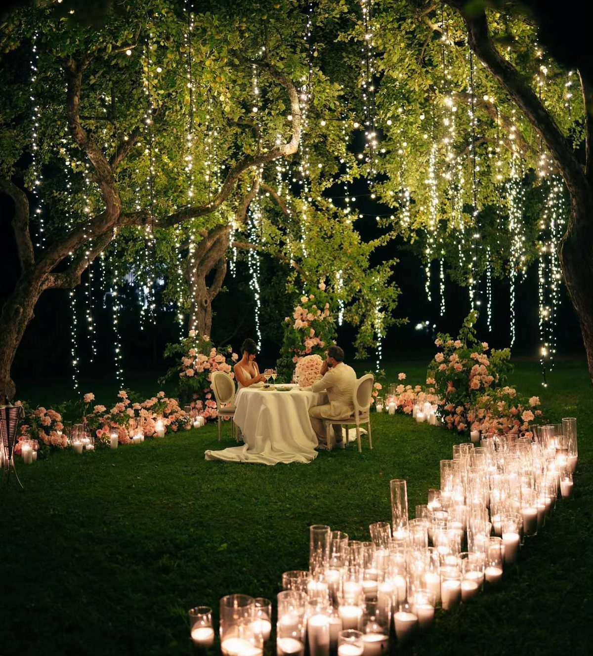 Strings of glowing fairy lights hang from tree branches over a romantic outdoor dinner setting.