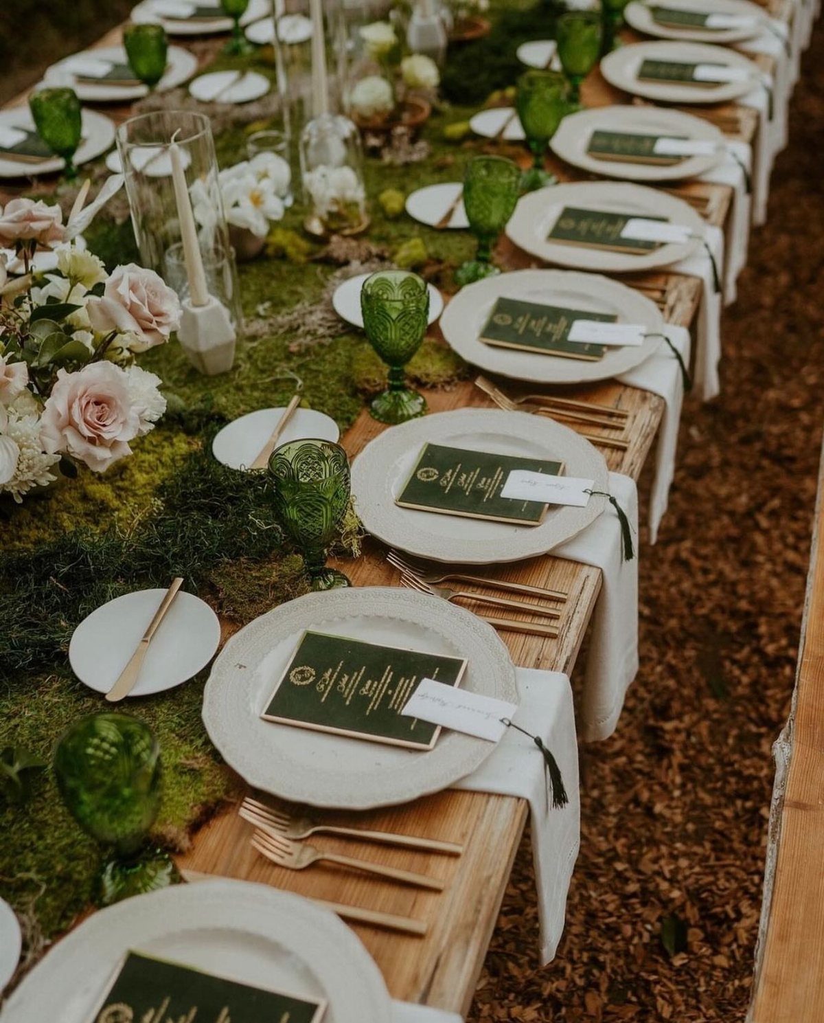 A long wooden wedding table decorated with a thick moss runner, white plates, and green glassware.