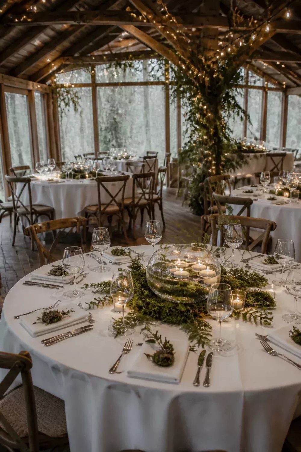 A round wedding table set with white linens, greenery garlands, and small candles in a rustic setting.